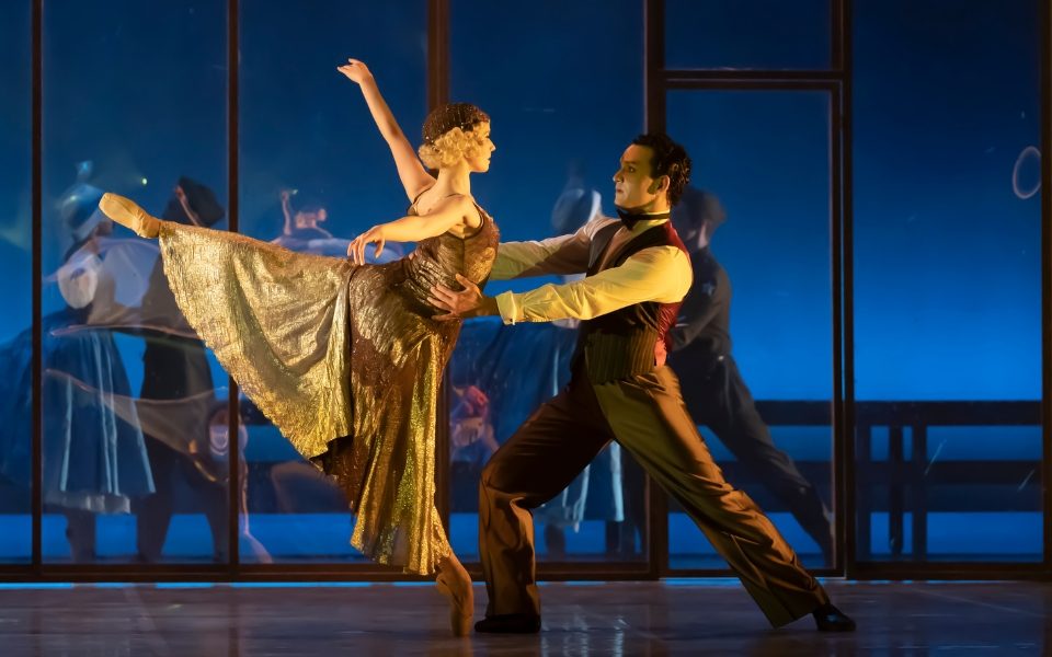 A man and woman in 1920s costumes facing each other and dancing together. The man holds the woman as she balances in a ballet post