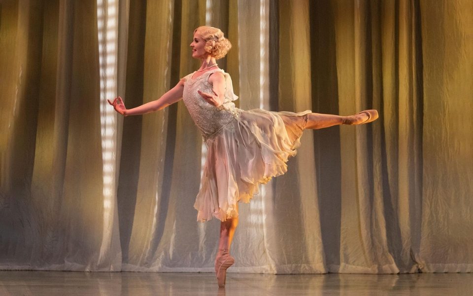 Dominique Larose as Daisy Buchanan in a white 1920s dress and curled blonde hair. She stands on pointe on one leg in a ballet pose.