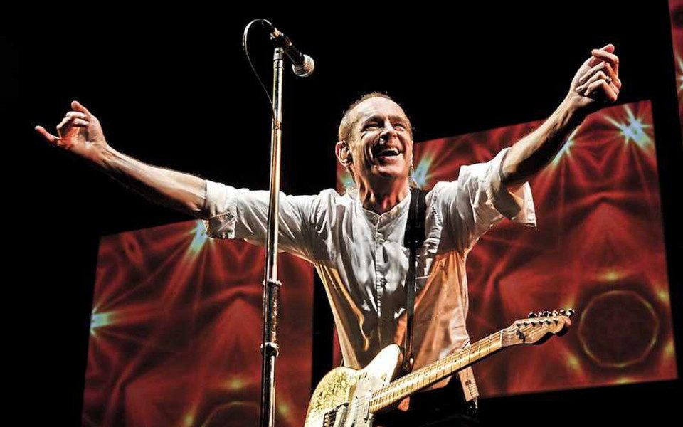Francis Rossi is on stage behind a microphone. His arms stretched toward the crowd and a guitar hangs from his neck. There are red and yellow images behind him on screens.