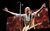 Francis Rossi in concert Francis Rossi is on stage behind a microphone. His arms stretched toward the crowd and a guitar hangs from his neck. There are red and yellow images behind him on screens.