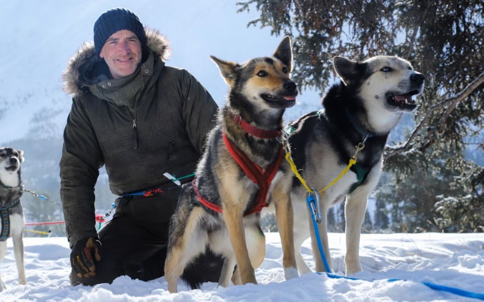 Gordon Buchanan in a winter coat and hat kneeling in the snow next to two dogs in harnesses