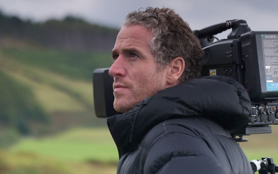 Gordon Buchanan in front of a large video camera staring out at the green hilly landscape