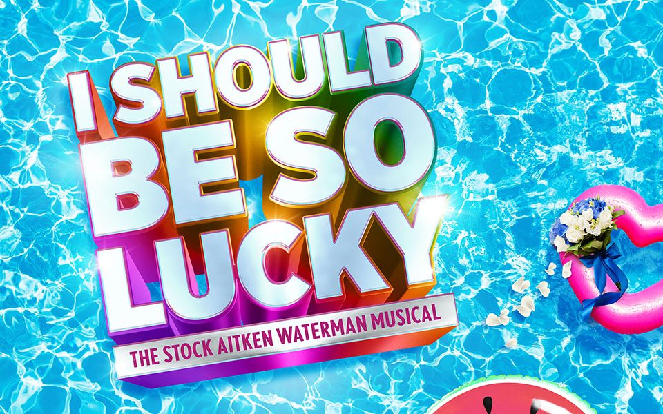 There are two pool floats, heart-shaped and watermelon-shaped, on the water. The I Should Be So Lucky logo is in the centre of the image in block letters with rainbow borders.
