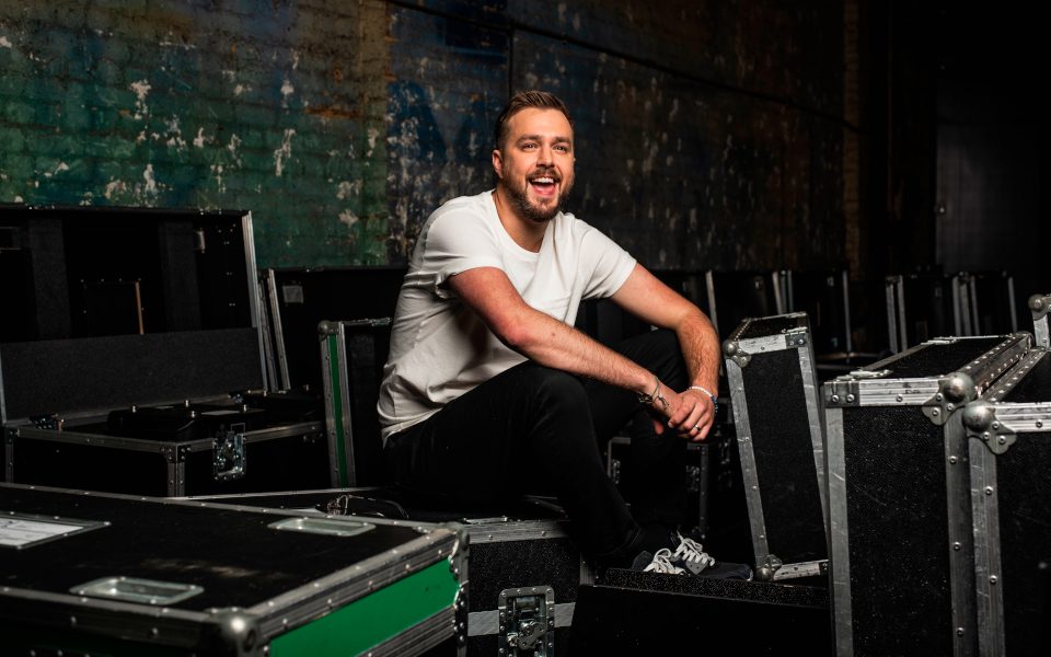 Iain Stirling has a big smile and is wearing a white shirt and black trousers sitting amongst the equipment boxes. The background is of colourful bricks.