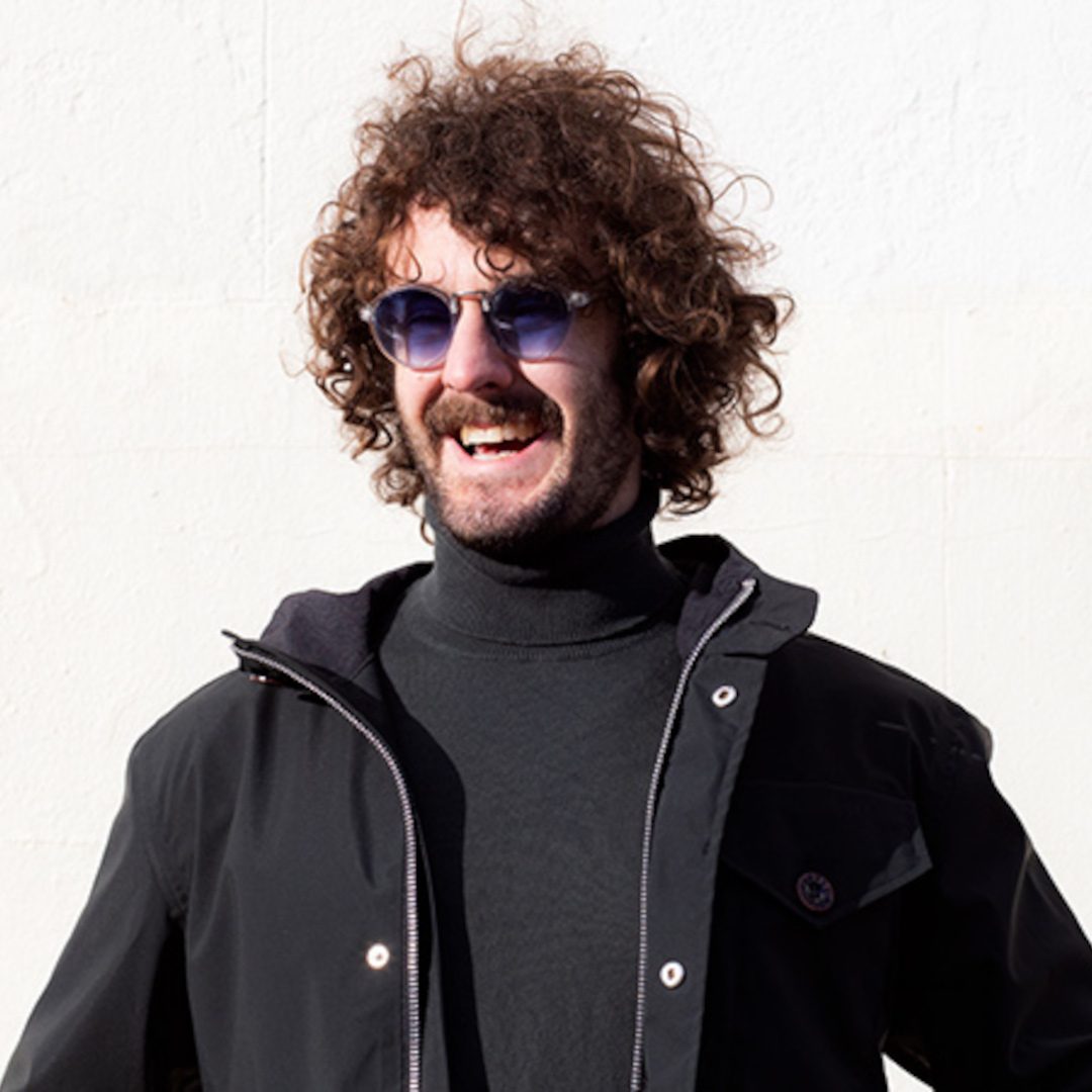Josh Pugh smiles as he wears a turtleneck jumper and black jacket. He is wearing blue sunglasses in front of a white wall.