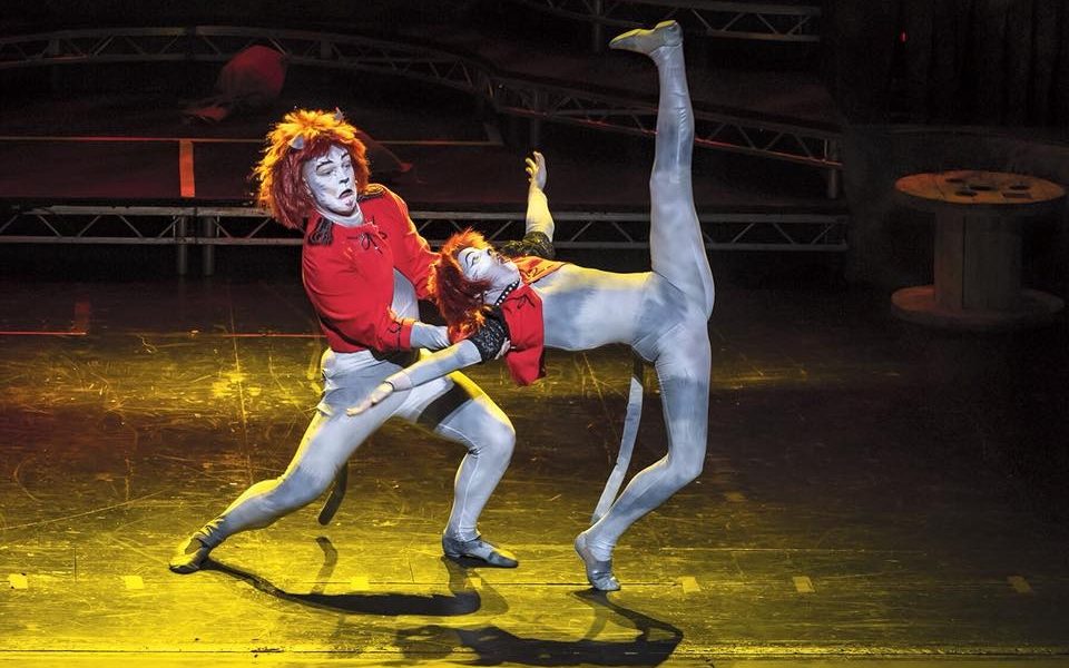 Two dancers dressed as grey cats with red jackets. One holds up the other as they extend their leg straight up in the air