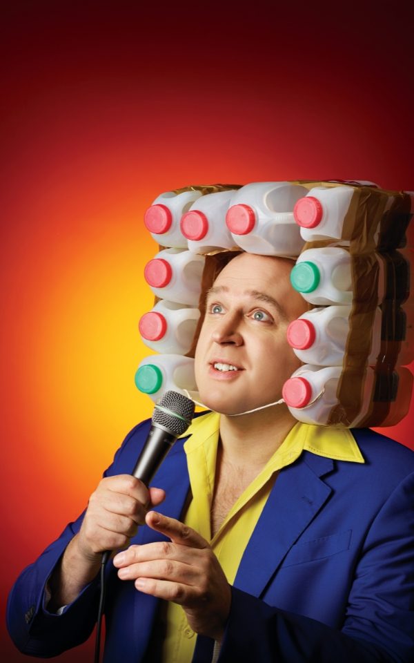 Tim Vine with milk bottles taped in a square around his head. He holds a microphone and looks off camera.