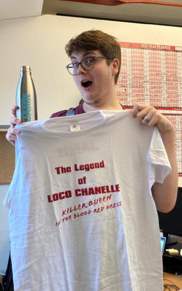 Paul is photographed with merchandise from Everybody's Talking About Jamie. Paul is a holding a water bottle with the logo and a white shirt with red writing on it that reads, 