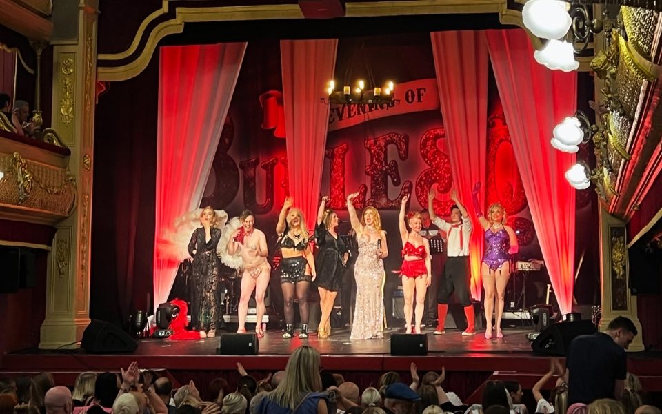 The cast of An Evening of Burlesque in front of a red backdrop on stage waving at a full audience at City Varieties