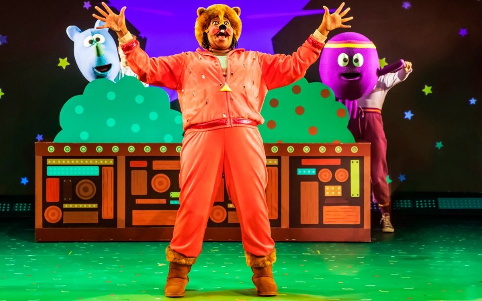 A person dressed as an animal in an orange jumpsuit and facepaint. There are two Hey Duggee puppets hiding in cartoon bushes in the background
