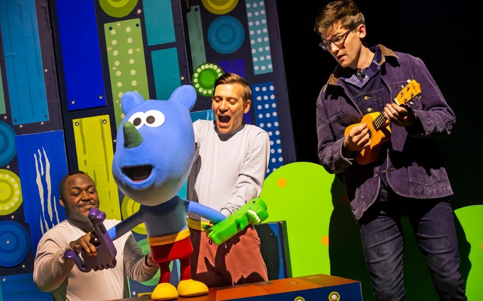 A blue rhino puppet standing on a table with a man playing the ukulele next to him