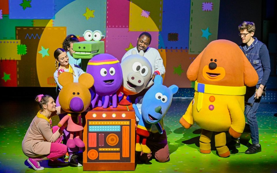 Colourful animal puppets with puppeteers gathered in a group with human-sized Duggee puppet (a dog in yellow jacket) next to them.