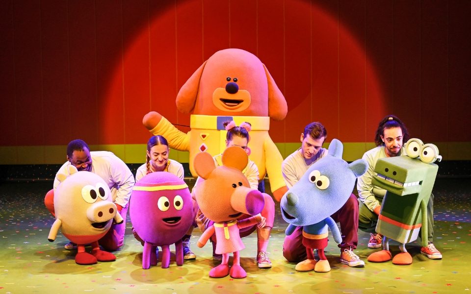 Large puppets of the colourful characters from Hey Duggee on stage in front of a red curtain. Duggee, a dog in a yellow coat, is in the middle.