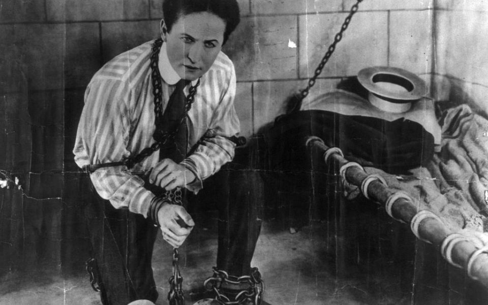A black and white photo of Harry Houdini crouching down and wrapped in chains with metal balls weighing him down. There is a note underneath saying 'Stone walls and chains do not make a prison - for Houdini'