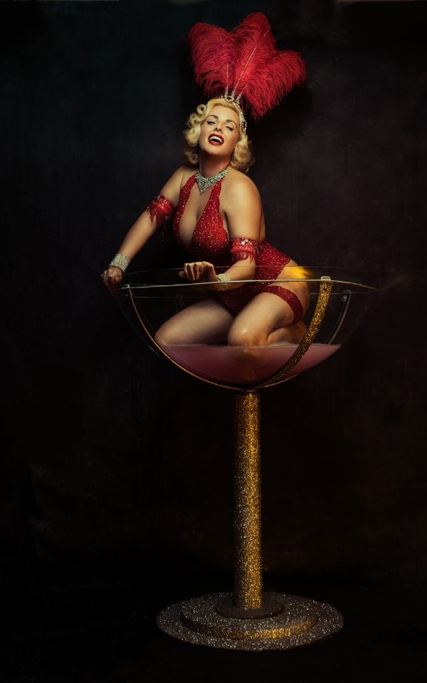 A woman in a red showgirl costume with red feathers in her blonde hair laughing at the camera. She crouches in a giant gold sparkly champagne coupe full of soapy water.