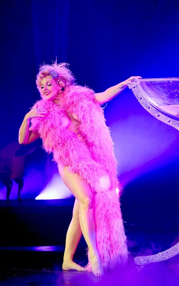 Isabella Bliss in a full length pink feather boa covering her body as she holds the side of a giant champagne coupe