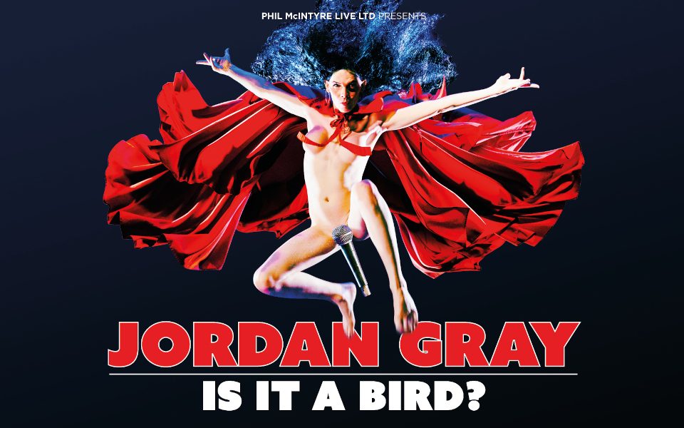 Jordan Gray is jumping in the air with long black hair and a red cape flowing behind. Jordan is nude with the string of the cape, and microphone strategically placed.