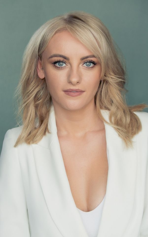 Katie McGlynn looks at the camera. She has long blonde hair and is wearing a white jacket.