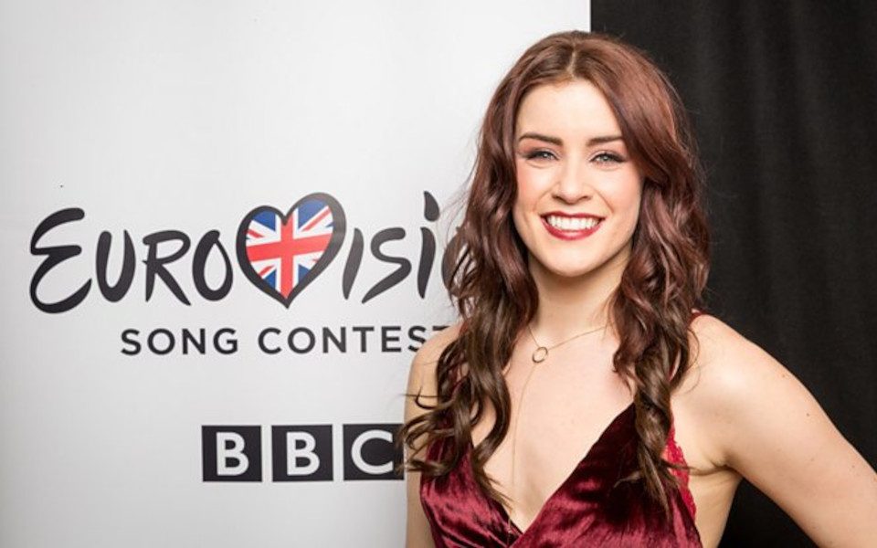 Lucy Jones is wearing a velvet red dress. She is in front of a Eurovision Song Contest poster.