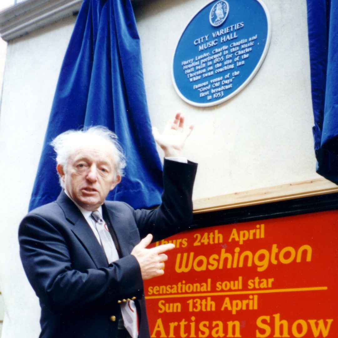 Paul Daniels in a suit pointing up at the Blue Plaque on the wall of City Varieties Music Hall