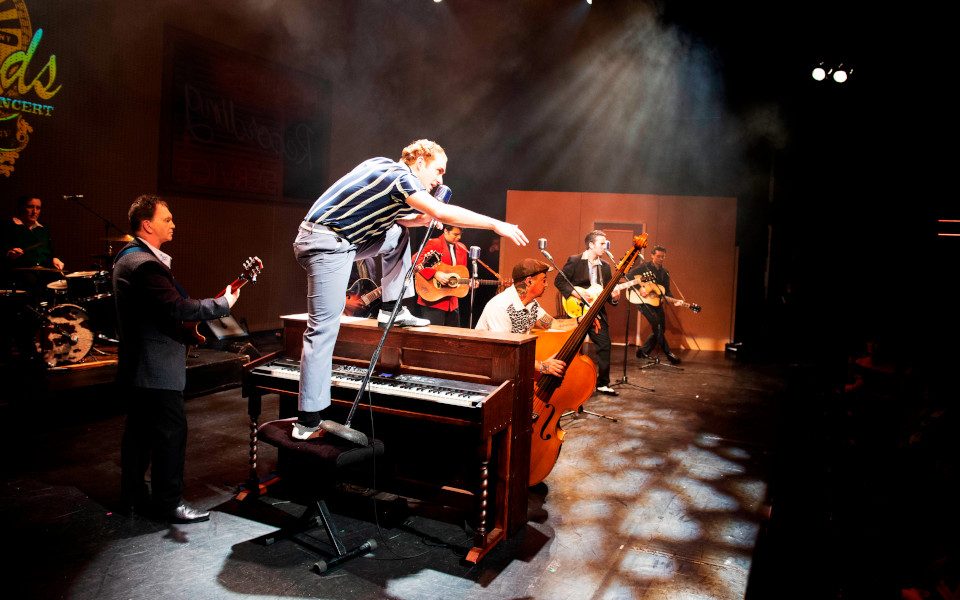 Musicians perform on stage. A man is standing on top of the piano singing into a mic.