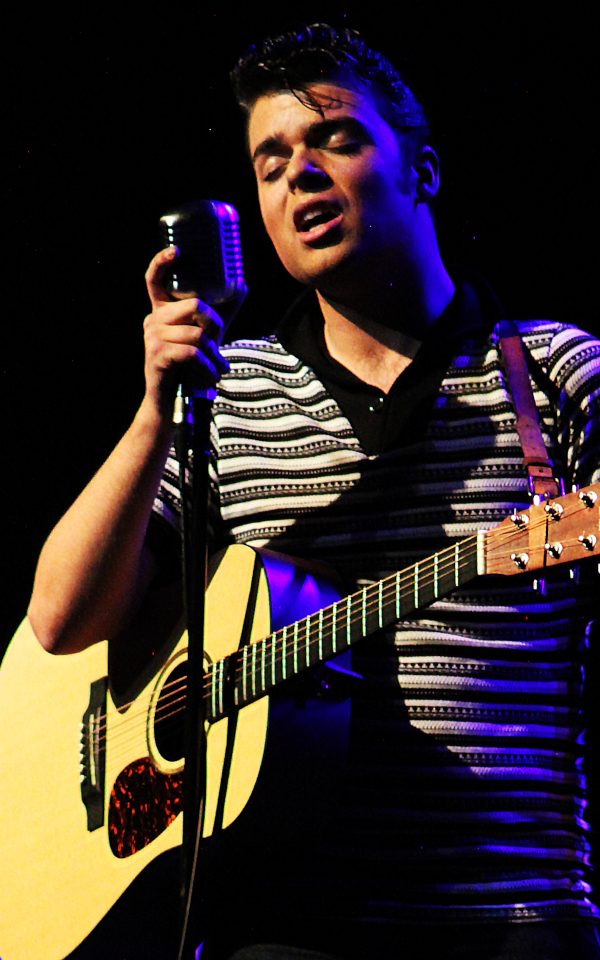 A singer performs in front of a standing mic with a guitar hanging around his neck.