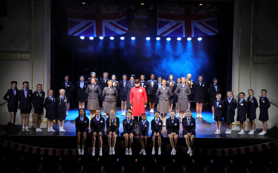 The D-Day Juniors sit on the edge of the stage in their 1940s-inspired uniforms, in front of Sergeant Major Colin Thackery in his red uniform and The D-Day Darlings in their 1940s-inspired uniforms around him.
