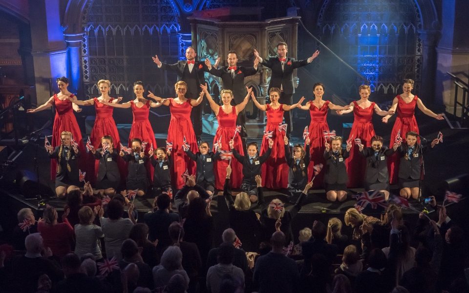 The D-Day Darlings in red dresses onstage with their arms out towards the audience. Behind them stand three men in suits with red bow ties (The Bombshell Beaus) and, in front of them, sit The D-Day Juniors in 1940s uniforms waving British flags.