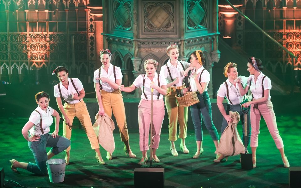 Eight women in different coloured pastel trousers with white shirts, braces and 1940s 'land girl' hair. They sing on stage and some carry hessian sacks, baskets, spades and buckets.