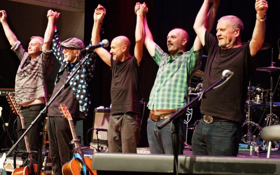 Members of Lindisfarne are standing on stage in the process of bowing at the end of a show. They are holding hands with all of their arms raised.