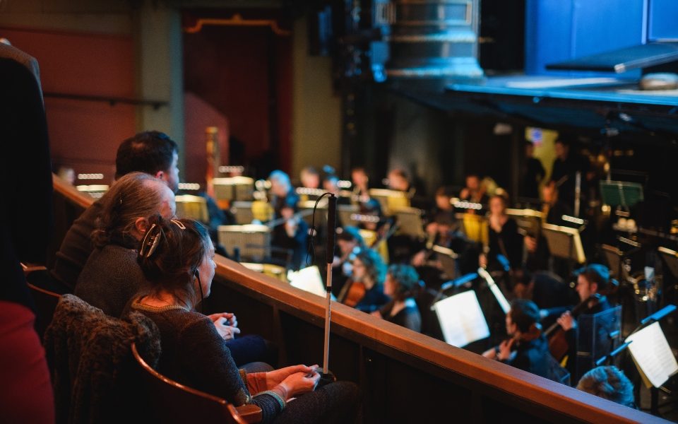 A woman with a mobility cane in the auditorium of Leeds Grand Theatre in front of the orchestra and stage.