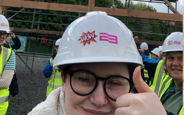 A selfie of Imogen Hinchcliffe doing a thumbs up in front of the building site, wearing a hard hat with WOW 23 on it.