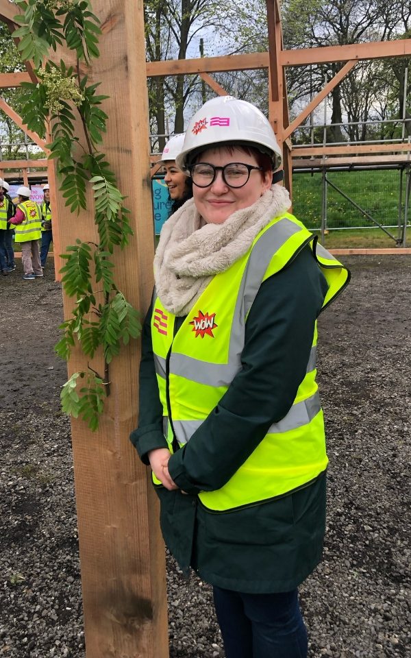 Imogen Hinchcliffe in a high vis jacket and white hard hat next to a wooden beam with leaves trailing off it