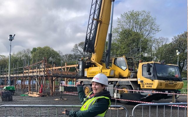 Imogen Hinchcliffe smiling and gesturing towards a big yellow crane