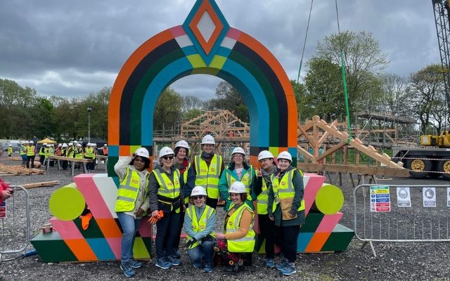 A group of women and non-binary people in high vis jackets and white hard hats smiling in front of a colourful arch