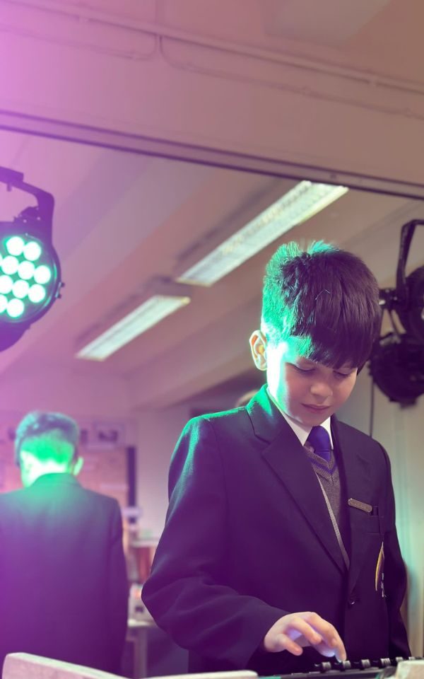 A student in a blazer and tie with short hair works with the lighting desk and rig, with his reflection caught in the mirror