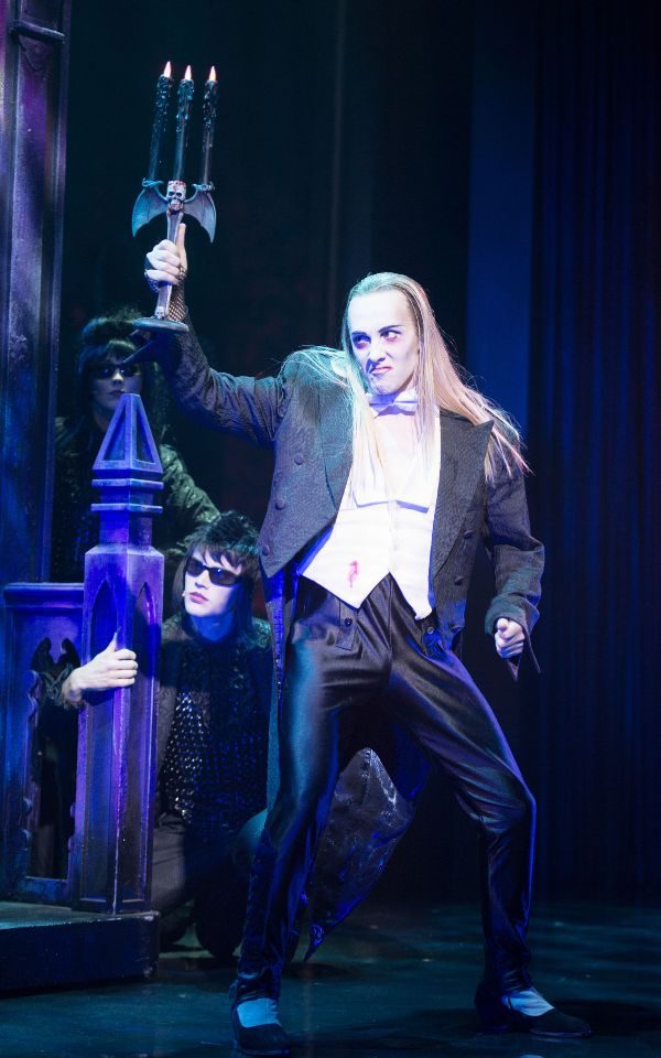 Dressed in a hunched tuxedo and holding a gothic black candelabra with black lit candles, Riff Raff from The Rocky Horror Show is in the foreground. In the background, a gothic character peers at them.