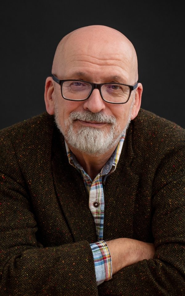 A headshot of Roddy Doyle looking at the camera with his arms folded. He is wearing a shirt and suit jacket and glasses.