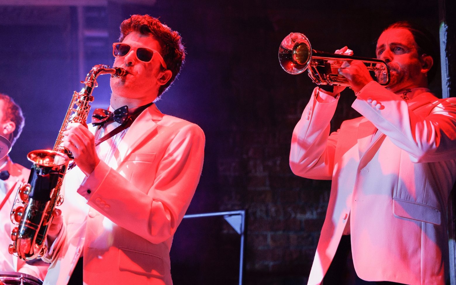 Two men in white suits and sunglasses. One plays the saxophone and the other plays the trumpet.