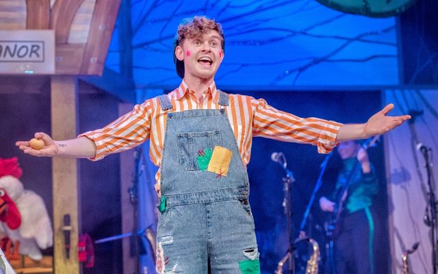 A man in a stripy shirt and denim overalls with his arms outstretched.