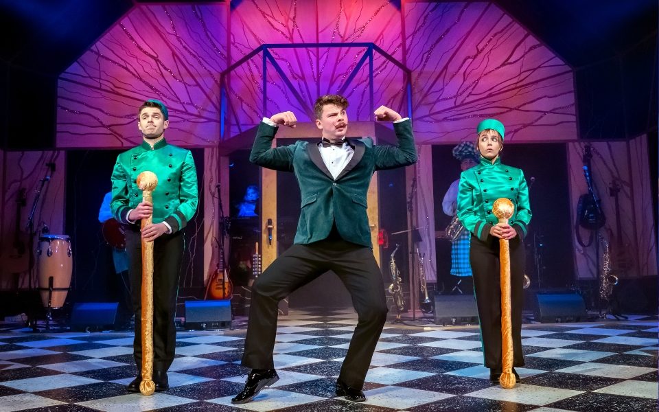 A man in a velvet suit posing in the middle of two servents in green costumes.