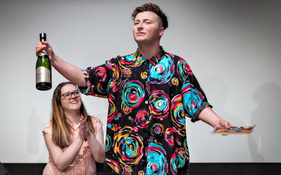 Bradley Thompson is giving a speech and wearing a colourful shirt and holding a package and bottle of prosecco. Another LAIT member is in the background clapping.