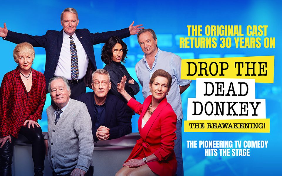 Over a blue backdrop, the cast of Drop The Dead Donkey pose in formal attire. The title reads 'Drop The Dead Donkey: The Reawakening' in yellow and black.