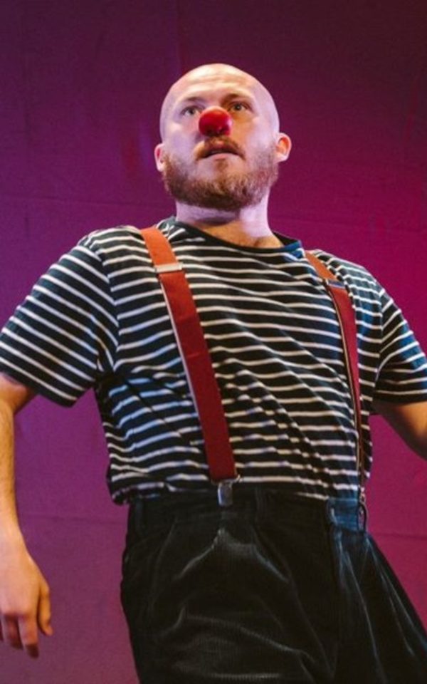 James Lewis-Knight is performing on a stage as a clown. He is wearing red suspenders and a red nose, accompanied by a striped shirt and black trousers in front of a colourful background.