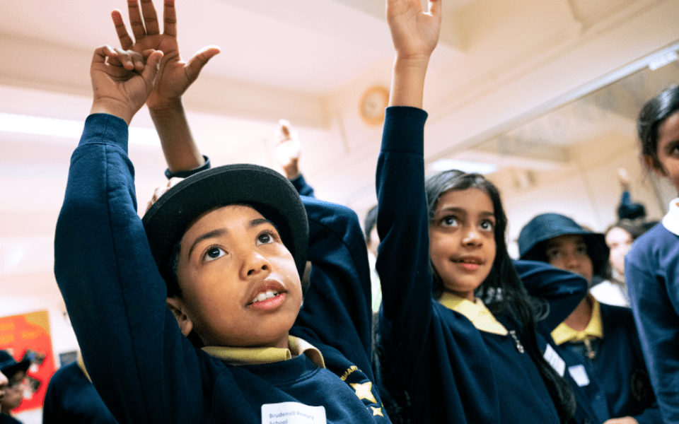 A pair of primary schoolchildren in costume hats put their hands up to answer a question