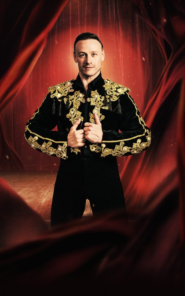Kevin Clifton in a black and gold Latin dancing costume in front of a red curtain
