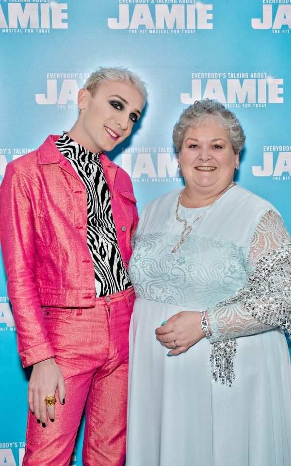 Jamie Campbell in a pink jacket and trousers with his mother Margaret Campbell in a pale blue dress smiling in front of the Everybody's Talking About Jamie press board.