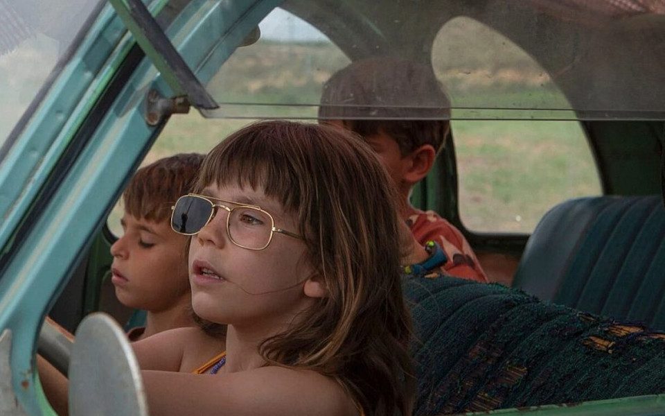 Three kids are in a car with one driving. The driver has sunglasses on with a lens missing. The window is opening and the seats are worn-out.