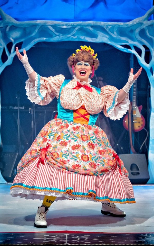 Simon Nock in a colourful maid costume as the panto dame. He stands in front of an icy woodland backdrop.