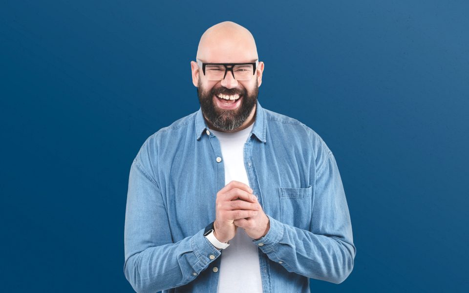 Tom Davis is clasping his hands and laughing. He is wearing a blue t-shirt and black glasses; he is standing in front of a blue background.